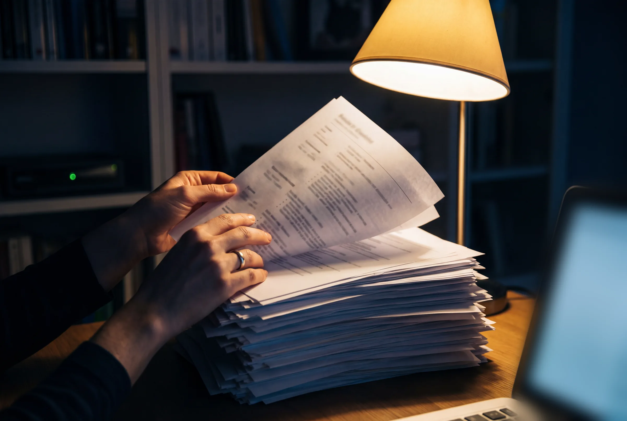 A tall stack of unread CVs under a desk lamp late at night, hands leafing through the pages.